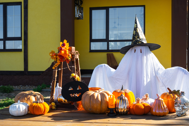 Child in bed sheet with slits like ghost in witch's hat and Halloween decor on the porch of the house outside in the yard of pumpkin, lantern, garlands, ack lantern. party, autumn mood