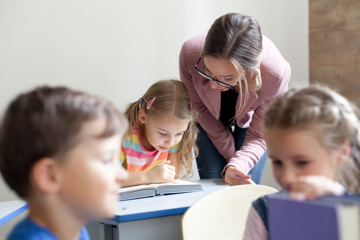 School teacher helping pupils studying, reading book. Lesson for kids in elementary college. Children sitting at desks in classroom. Woman guiding in-person, teaching primary students. Back to school