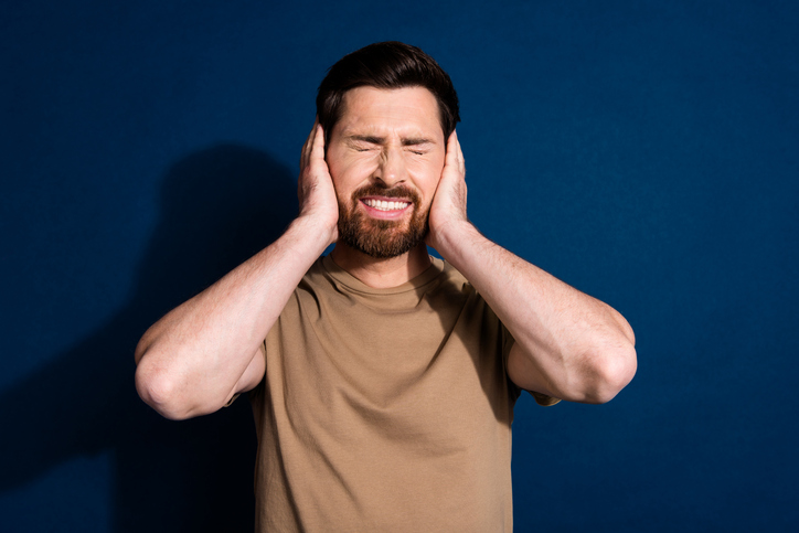 Photo of offended man with stylish beard dressed beige t-shirt hold hands on ears ignore loud noise isolated on dark blue color background