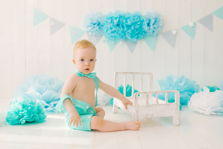 A one-year-old boy in a summer suit with a butterfly sits on the photo zone of his birthday in blue colors with a baby cot