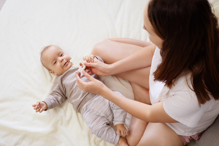 A mother is joyfully playing with her baby on a soft blanket at home. The room is filled with warmth, creating a nurturing environment for their interaction