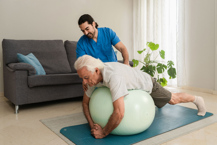 Male Physiotherapist assisting senior man doing exercises with fitness ball at home