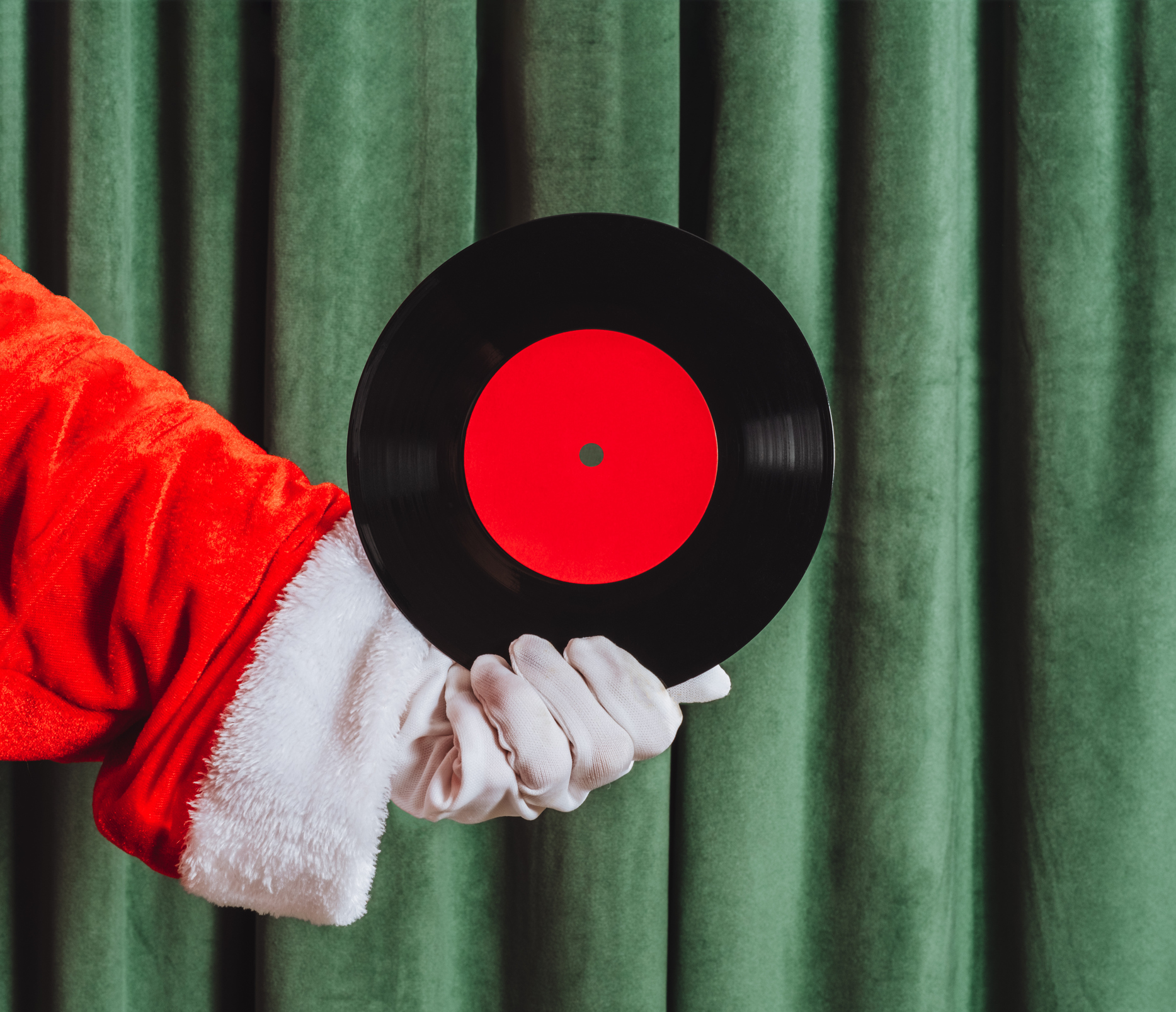 Santa Claus hand in red glove holding a vinyl record with red label. Retro holiday music concept with green curtain background.
